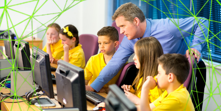 a male teacher in blue shirt lent over at a desk helping school kids wearing yellow polo tops do something on the computer.
